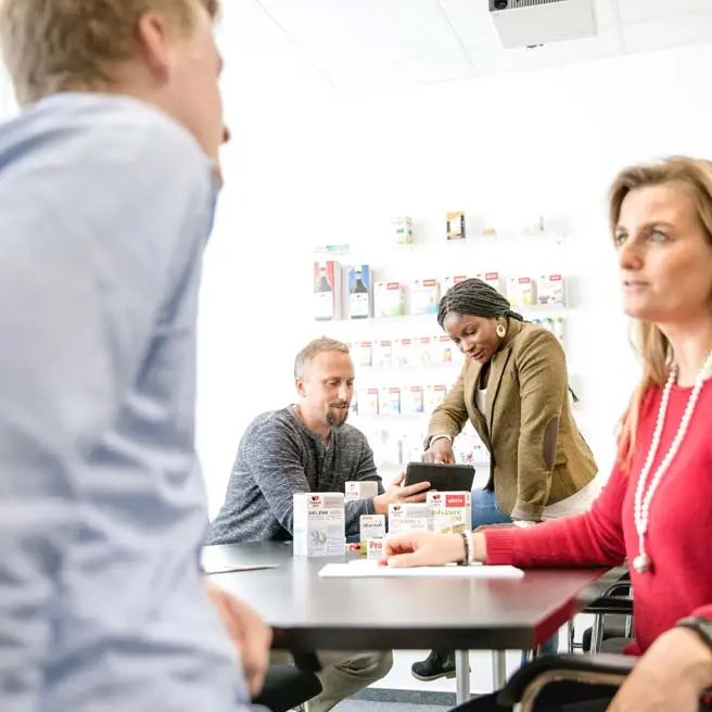 Four people are sitting around a table in a meeting room. One person is holding a tablet while another examines a product box. Various product packages are displayed on a shelf in the background, indicating a discussion about these items.